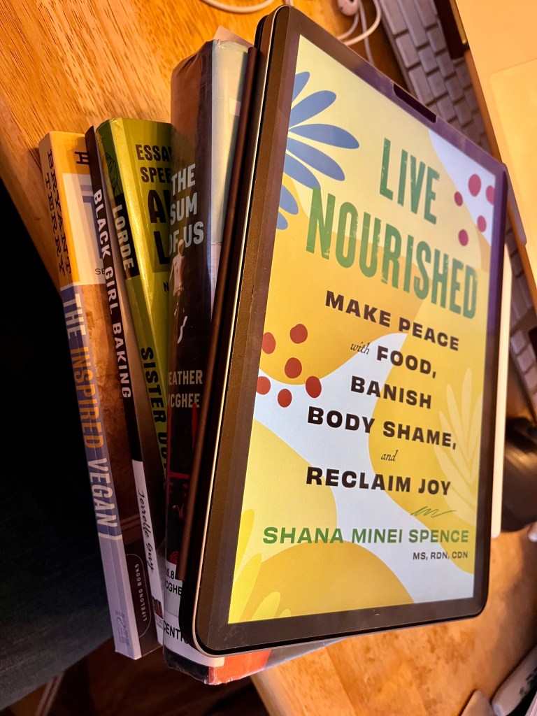 Photo of an iPad with a Kindle book, a library book, a book of essays, and two cookbooks.